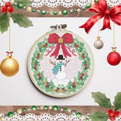 a cross stitch christmas ornament hanging from a wooden frame with holly wreaths and baubles