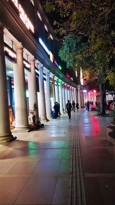 people are walking down the sidewalk in front of some tall white pillars at night time