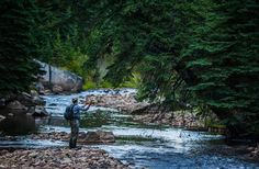 a man standing in the middle of a river holding a fishing rod and wearing a backpack