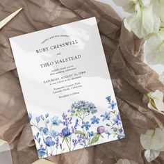 a wedding card with blue flowers on it next to some white and purple flowers in the background