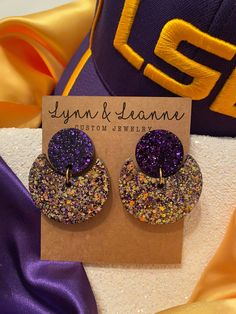 purple and gold glittered earrings on top of a card next to a baseball cap