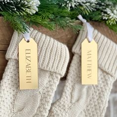 two christmas stockings with tags hanging from them on a wooden table next to evergreen branches