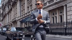 a man in a suit and sunglasses walking down the street next to a black car