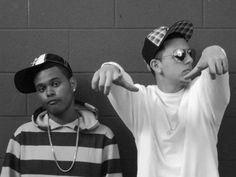 two young men standing next to each other in front of a brick wall holding their hands up