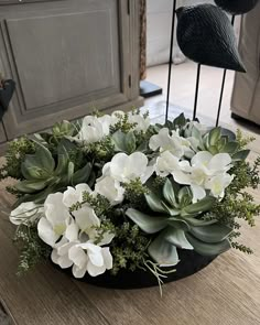 white flowers and greenery are arranged in a black bowl on a table next to a bird statue