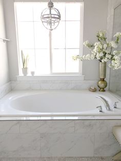 a white bath tub sitting under a window next to a vase with flowers in it