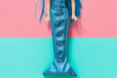 a mermaid doll with blue hair standing next to a pink and turquoise wall