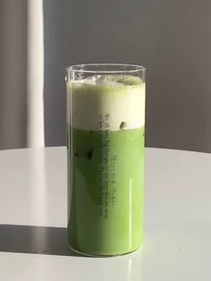 a glass filled with green liquid sitting on top of a white table next to a window