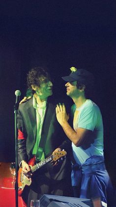 two men standing next to each other in front of a microphone, one holding a guitar