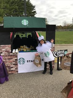 a woman dressed in costume standing next to a table with starbucks bags on it and holding up her hand