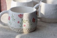 two white mugs sitting on top of a table next to each other with flowers painted on them