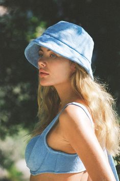 a woman wearing a blue hat standing in front of some trees and bushes with her hand on her hip