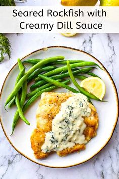 a white plate topped with chicken covered in creamy dill sauce next to green beans and lemon wedges