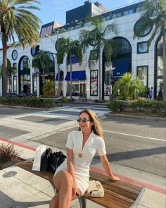 a woman sitting on top of a wooden bench next to palm trees in front of a building