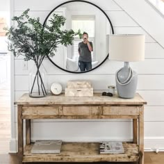 a person taking a selfie in the mirror above a table with a vase and lamp on it