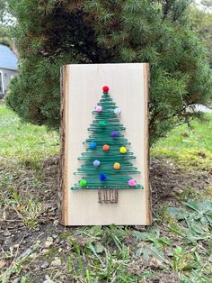 a small christmas tree made out of yarn on a wooden board in front of some trees