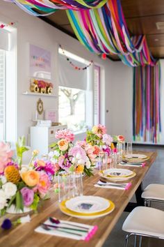 a long table with plates and flowers on it in front of colorful streamers hanging from the ceiling