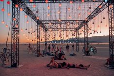 some people are laying on the sand under an open structure with lights hanging from it