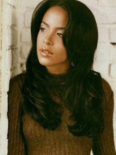 a woman with long dark hair standing in front of a brick wall wearing a brown sweater