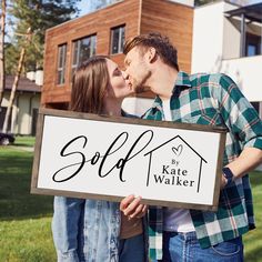 a man and woman kissing in front of a house holding a sign that says sold by kate walker