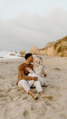 a man and woman are sitting on the sand at the beach, hugging each other