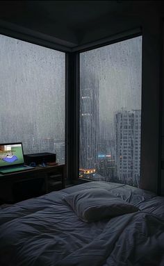 a bed in front of two large windows with rain falling down on it and a laptop computer sitting on top of the bed