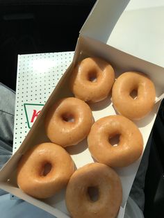 six glazed doughnuts in a box on the ground next to a person's lap