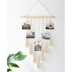 a white wall hanging with pictures and tassels attached to it, next to a potted plant