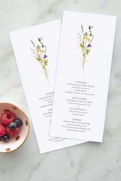 two cards with flowers and berries on them next to a bowl of raspberries