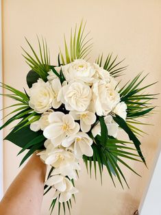 a person holding a bouquet of white flowers in their hand with palm leaves on the wall behind them