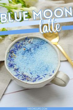 a white cup filled with blue liquid next to a gold spoon and flowers on a table