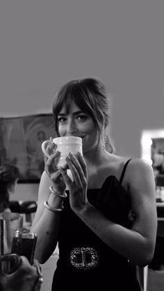 a woman in a black dress drinking from a coffee cup while another woman looks on