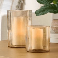 two glass candles sitting on top of a wooden table next to a potted plant