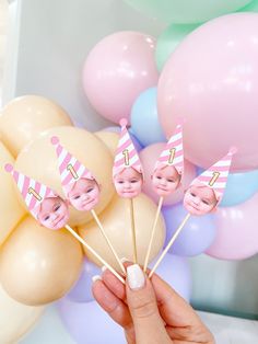 a person holding up some pink and white cake pops with faces on them in front of balloons