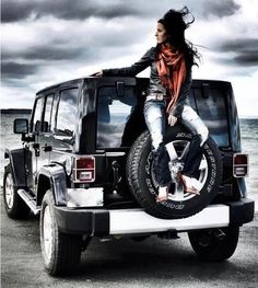 a woman sitting on the back of a black jeep parked in front of the ocean