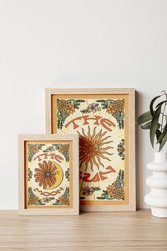 two framed pictures sitting on top of a wooden table next to a vase and plant