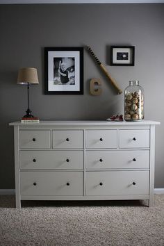 a white dresser topped with lots of drawers next to a lamp and pictures on the wall