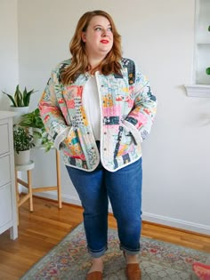 a woman standing in front of a white wall wearing blue jeans and a colorful jacket