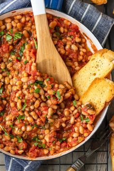 baked beans with tomatoes and parsley in a white bowl on a blue towel next to breadsticks