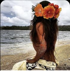 a woman with flowers in her hair sitting on the beach looking out at the water