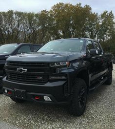 two black chevrolet trucks are parked in a gravel lot with trees and bushes behind them