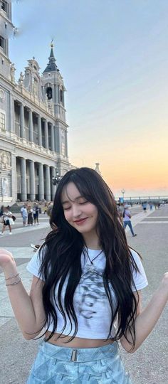 a girl with long black hair standing in front of a building and holding her hands out