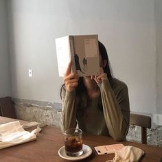 a woman sitting at a table with an apple box on her head and holding up a book in front of her face