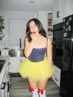 a woman in a yellow tutu skirt eating an apple while standing in a kitchen