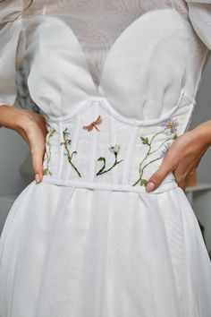 a woman wearing a white dress with embroidered flowers on the waist and sleeves, holding her hands in her pockets