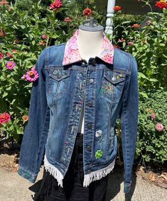 a blue jean jacket with patches on the back and pink flowered collar, sitting on a mannequin in front of some flowers