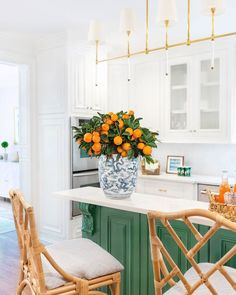 a vase with oranges on top of a kitchen island next to two wicker chairs