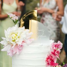 a white cake with flowers on top and people standing around in the background looking at it