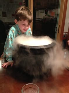 a young boy sitting at a table in front of a large pot with steam coming out of it