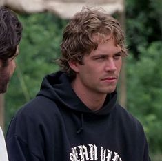 two men standing next to each other in front of some trees and one is wearing a black hoodie